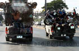 Auf dem Bild aus Thailand erkennt man ein Pickup Auto wo die Ladefläche mit Mopeds voll ausgenutzt ist. Auf der Pritsche beim Lastwagen links im Bild transportiert man Textilien und auf den Stoffballen sitzen noch drei Thailänder.